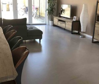 Grijze gietvloer in een woonkamer met hoekbank, eettafel en openslaande tuindeuren