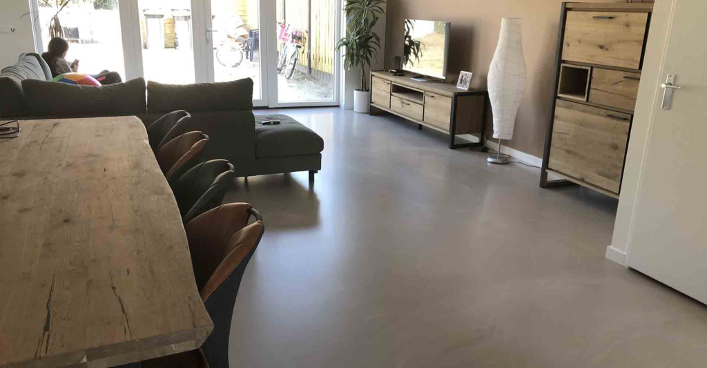 Grijze gietvloer in een woonkamer met hoekbank, eettafel en openslaande tuindeuren