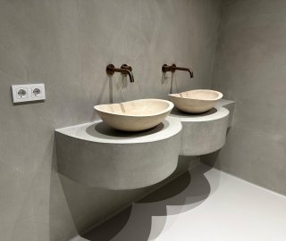 Zwevende microcement wastafel met twee travertijn waskommen en bronzen kranen